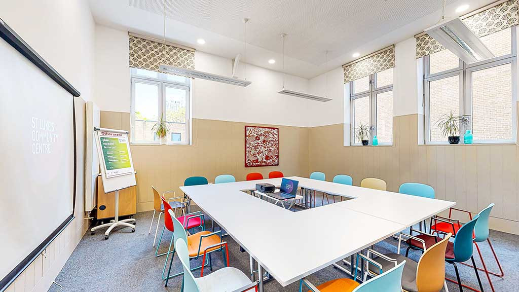 A community centre meeting room laid out in a boardroom square with colourful chairs, a projector screen and a flipchart
