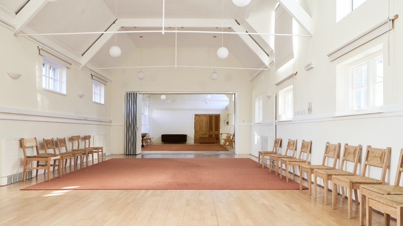 A bright community hall with a wooden floor, wooden chairs set around the edge and folding partition doors open to a second room